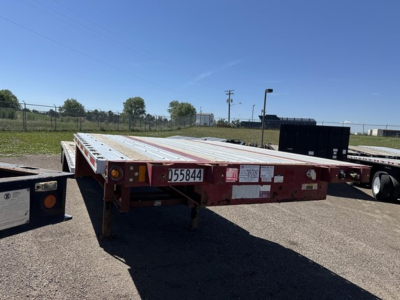 Flatbed trailer with drop deck in a parking lot showing side rails and rear frame; visible stickers and serial plate.