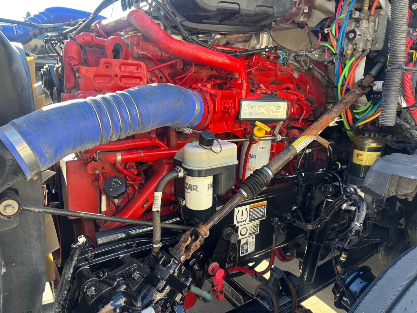 Kenworth T680 engine bay with red engine block, blue air intake hose, and various wiring and fluid reservoirs visible.