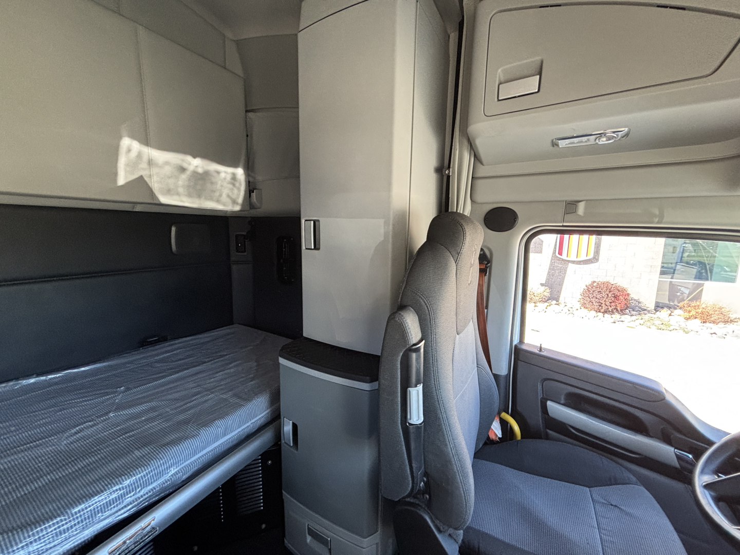 Kenworth T680 cab interior showing sleeper berth with single bunk, gray upholstery, overhead storage, and driver seat; visible window and dashboard area.