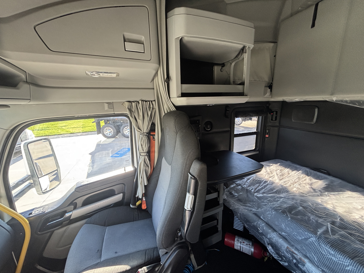 Kenworth T680 truck cab interior showing sleeper area with overhead storage, table, and passenger seat; plastic-wrapped bed and window view.