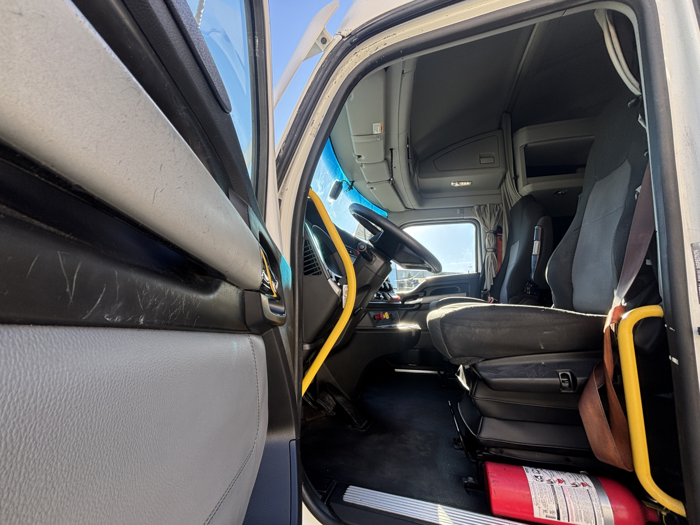 Kenworth T680 cab interior view from open door showing driver seat and passenger seat, yellow grab handle, dashboard, and steering wheel.