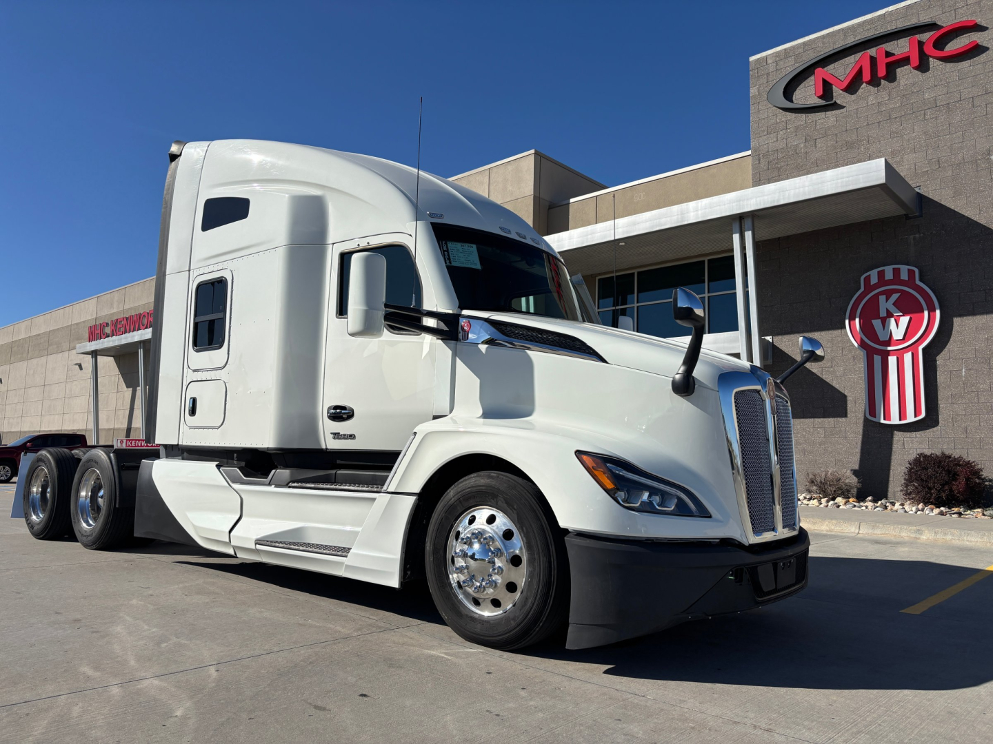 Kenworth T680 tractor unit in white, side view, with MHC/KW dealer building in background. No plate visible.