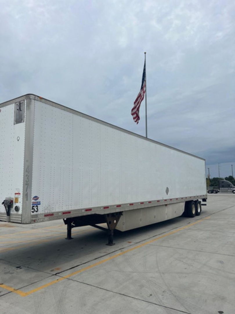 Utility van trailer showing side outline and front corner with leg support and flag in background.