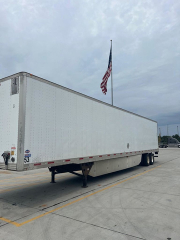 Utility van trailer showing side outline and front corner with leg support and flag in background.