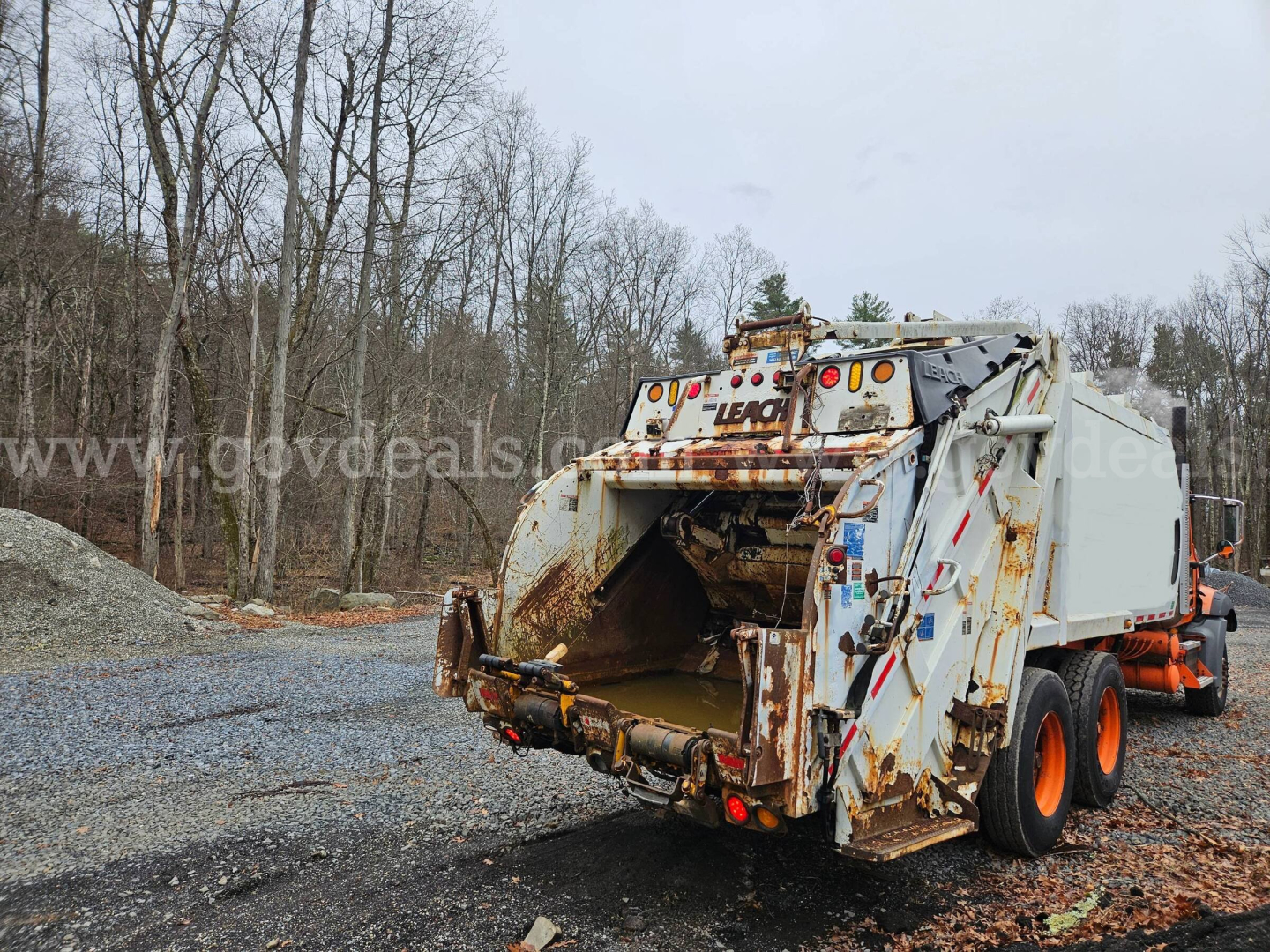 Rear view of a 2009 Mack GU813 refuse truck body with rusted hopper and orange wheels, showing rear loading chute and warning lights.