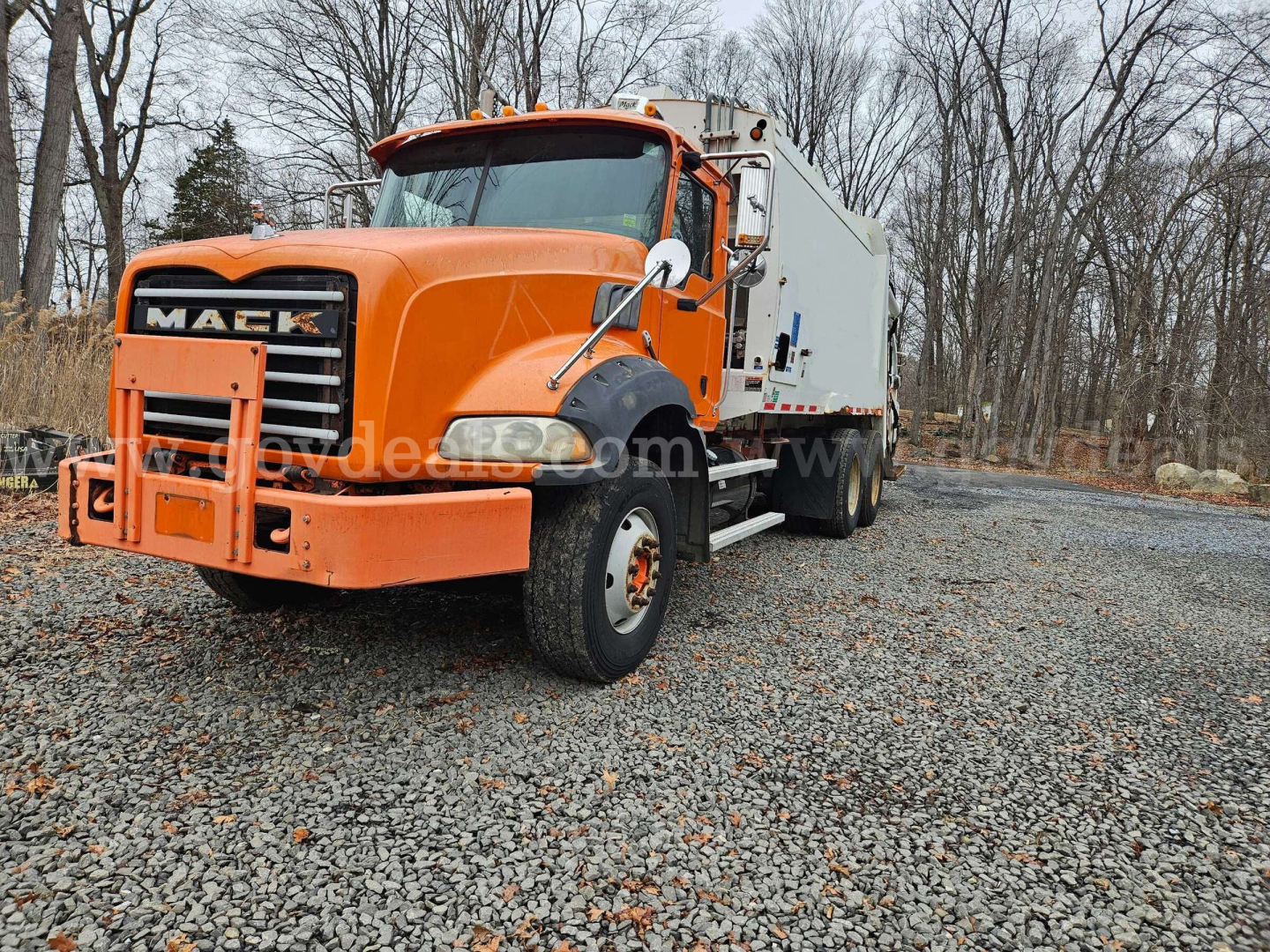 Mack GU813 heavy-duty truck in orange with box cargo bed, front grille, and dual rear wheels, parked on gravel with trees in background.