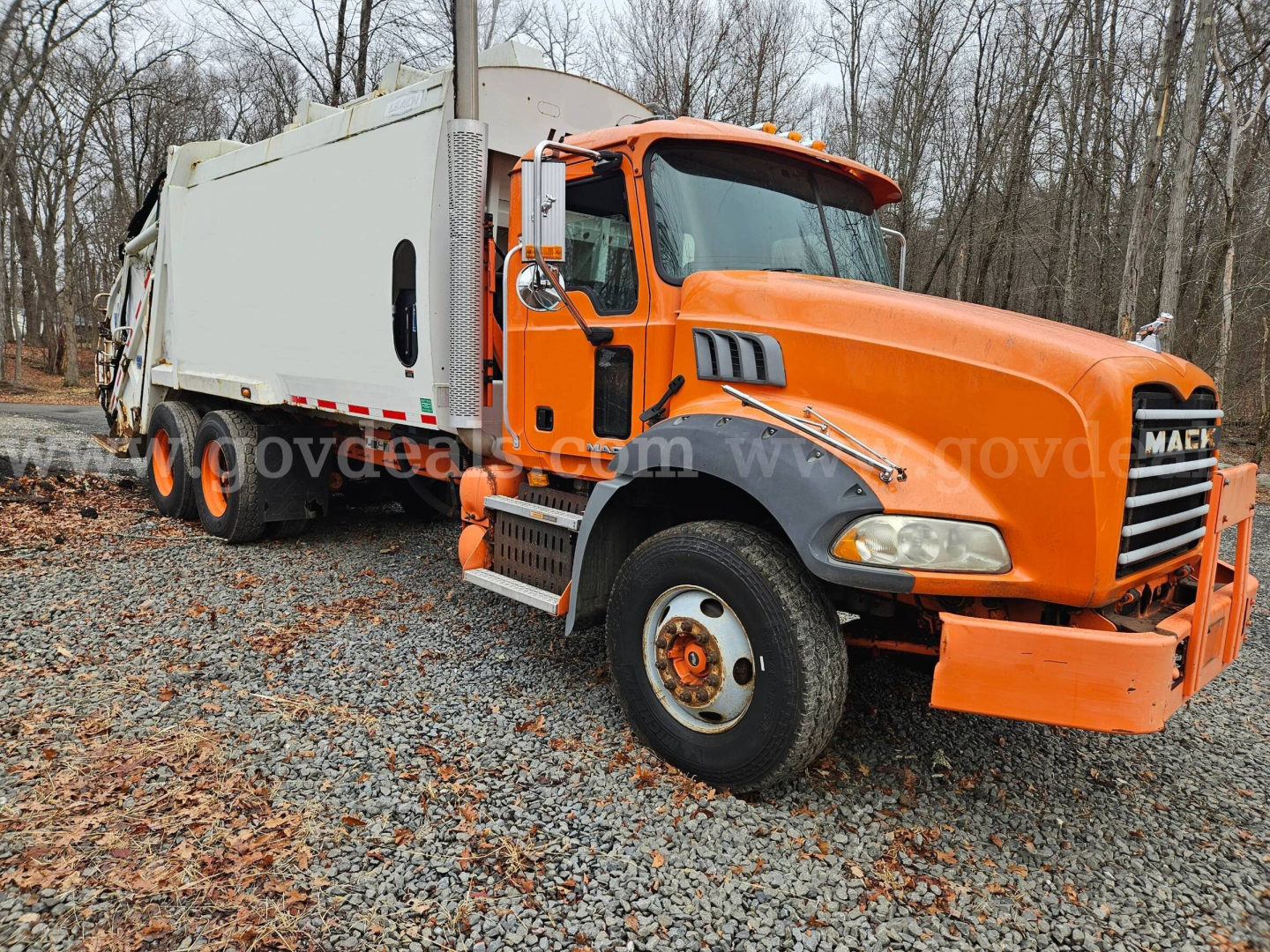 Mack GU813 truck in service with orange cab and gray refuse body, front-left view showing Mack grille, wheel, and side mirrors.
