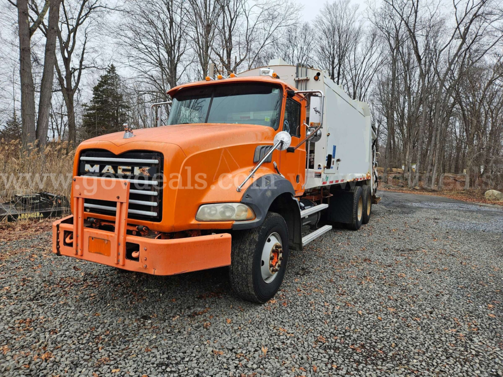 Mack GU813 truck in orange cab with front grille and utility body, visible VIN plate area and stock insignia in a gravel lot.