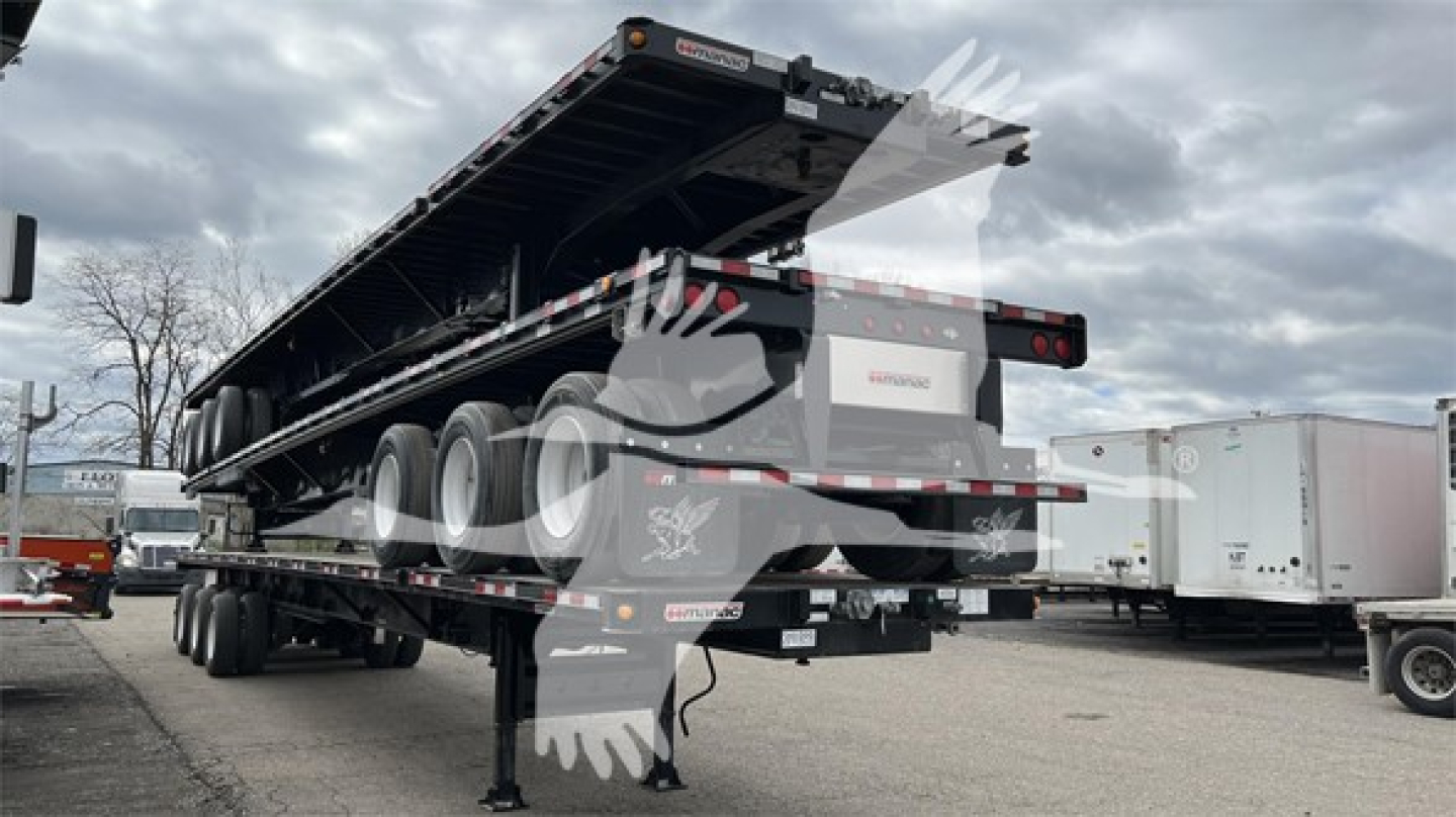 Manac extendable flatbed trailer viewed from rear-left, with multiple axles and a raised bed, loading ramps folded, warning labels and reflective tape visible.