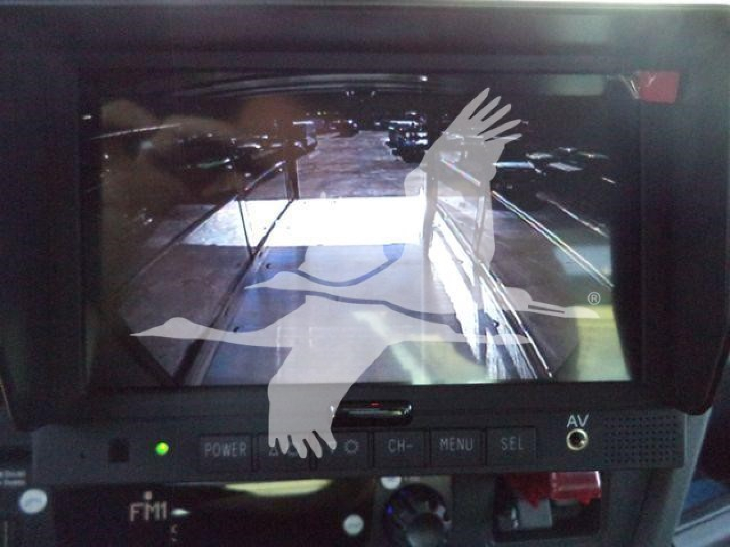 In-vehicle display screen showing a rearview camera feed with a reflection of a moving vehicle and logo watermark overlay.