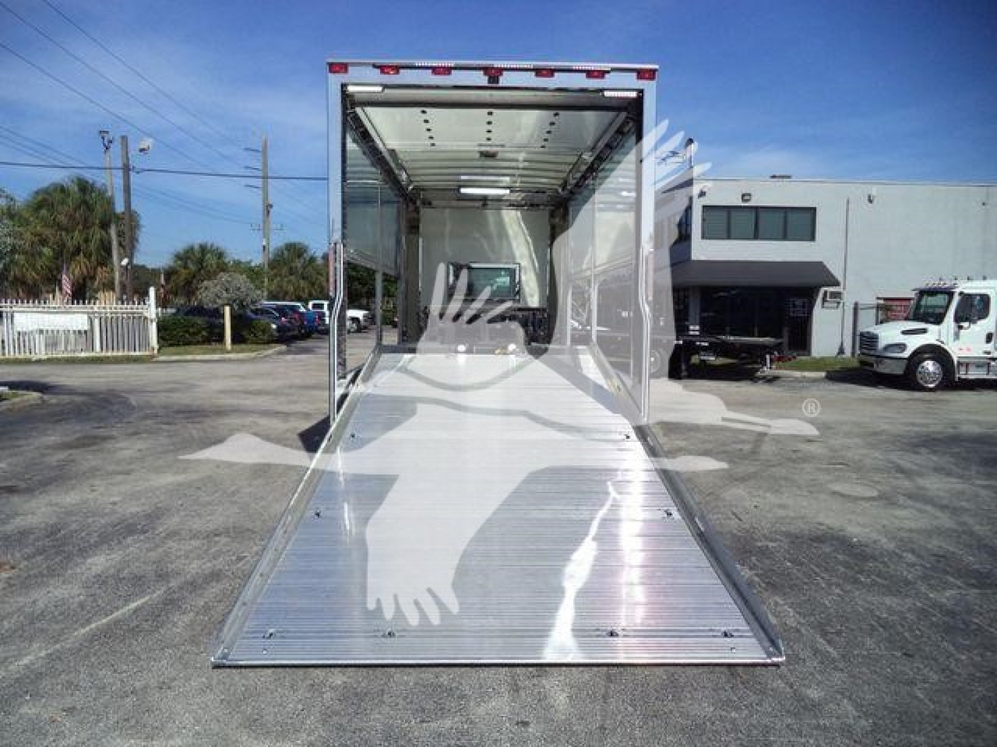 Rear view of a new 2025 Kenworth T280 rollback truck with an open aluminum ramp extending from the back, empty cargo area, and a dealership lot in the background.