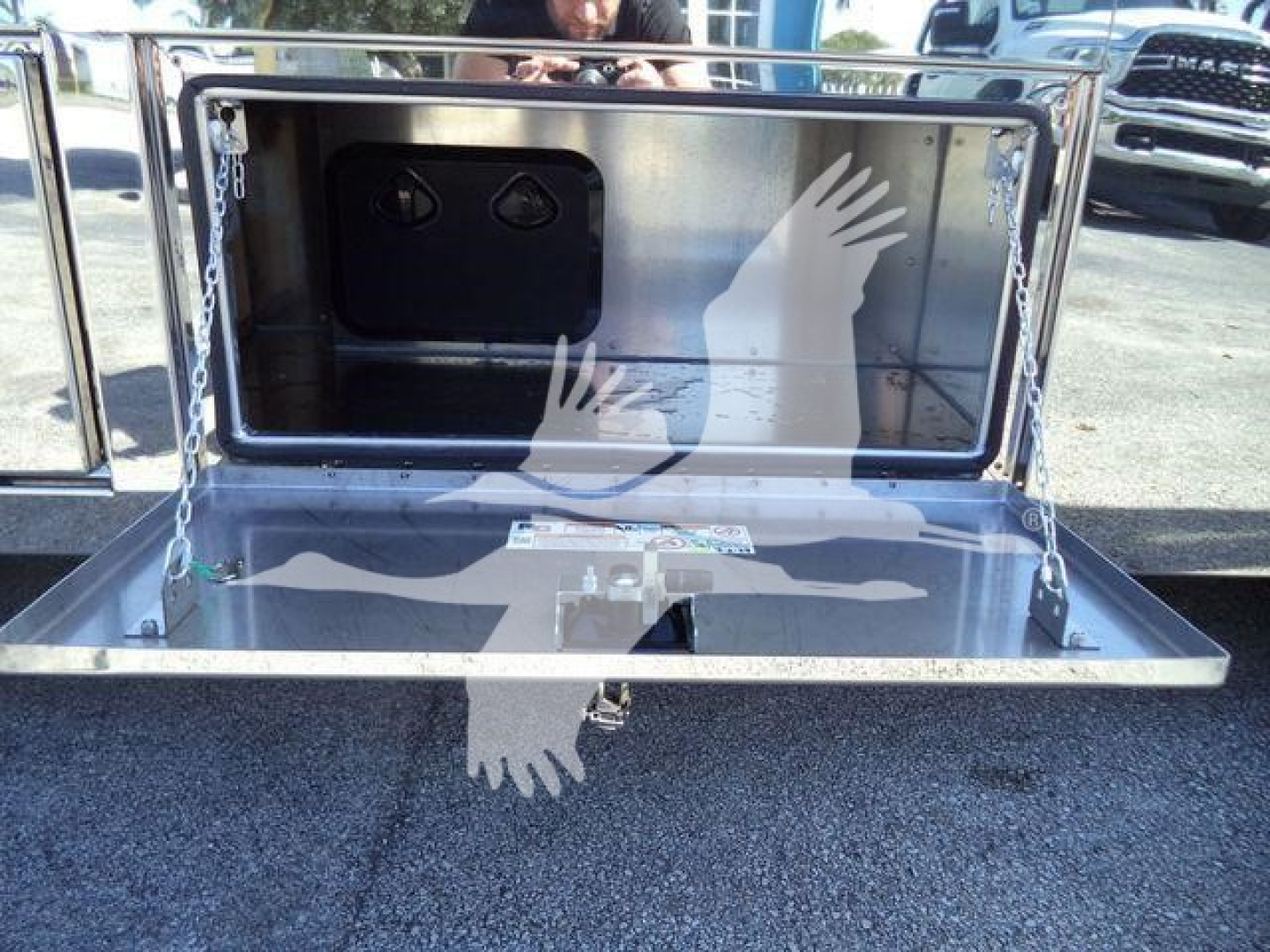 Truck equipment image showing a metal storage box with a hinged drop-down door and chain supports; interior has two circular fittings and a rectangular black panel.
