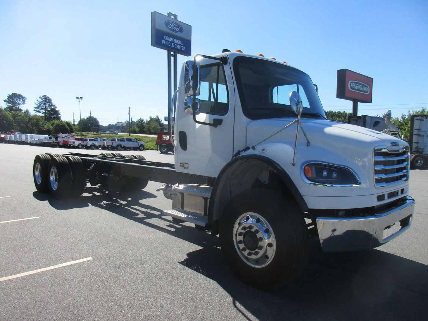Freightliner M2 106 Plus cab and chassis truck in a dealership lot showing front and side view with empty flatbed frame.