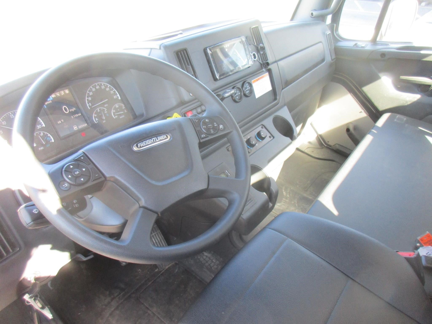 Cab interior of a Freightliner M2 106 Plus showing steering wheel with Freightliner badge, instrument cluster, center dash with touchscreen, and passenger seat.