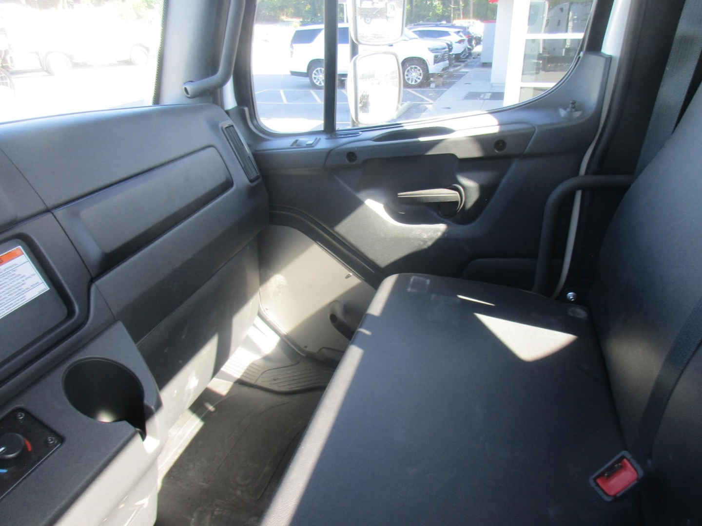 Freightliner M2 106 Plus cab interior with bench seat and side door panel, view from passenger side showing window, dashboard, and seat belt buckle.
