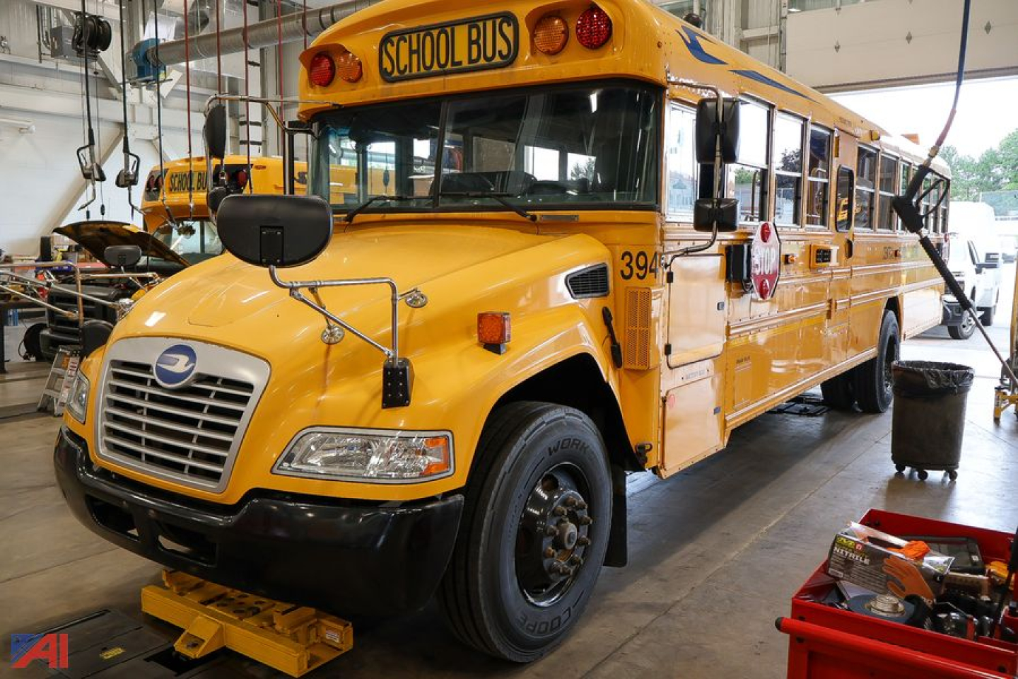 School bus showing front and passenger side in a workshop with yellow body, black bumper, and 'SCHOOL BUS' sign on roof; number 394 on door; wheel removed on front right; repair tools nearby.