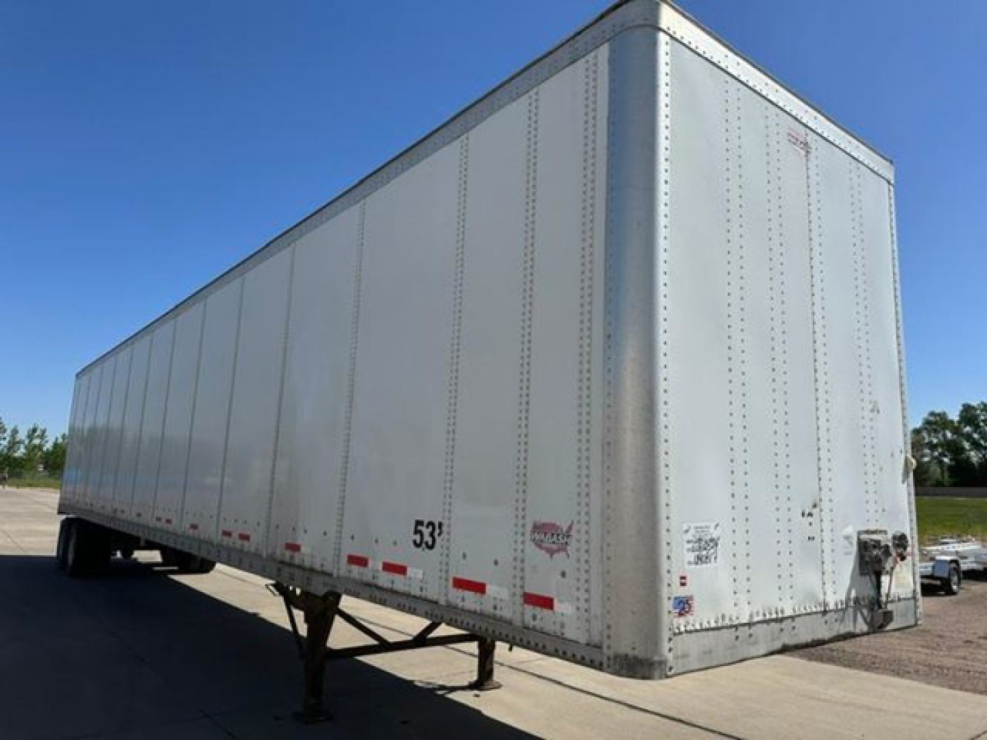 Trailer side and front corner showing a large white Wabash enclosed trailer with tandem axles, 53-foot length, riveted aluminum panels, rear doors, and side height markings.