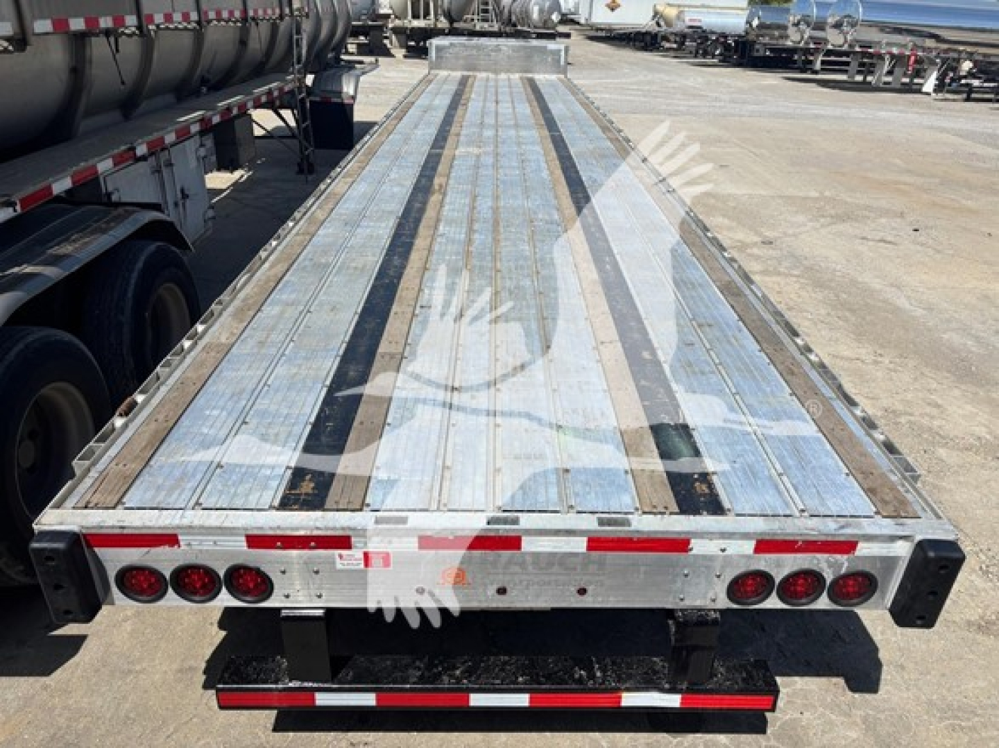 Flatbed trailer deck with metal planks and side rails, rear view showing tail lights and underride guard on a Transcraft trailer.