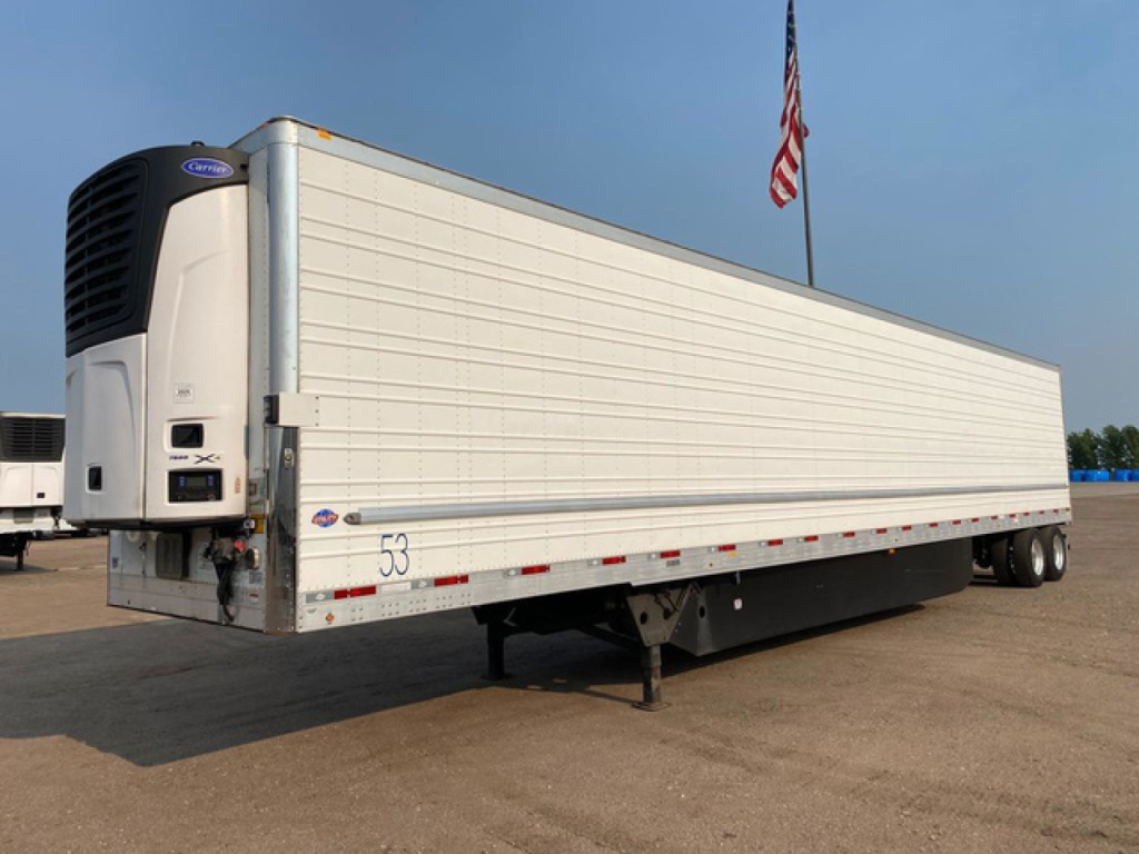 Trailer side with reefer unit at front left, white refrigerated trailer with blue trim and a Carrier cooling unit mounted at front. Visible text includes numbers on the side and the front header. American flag in background.