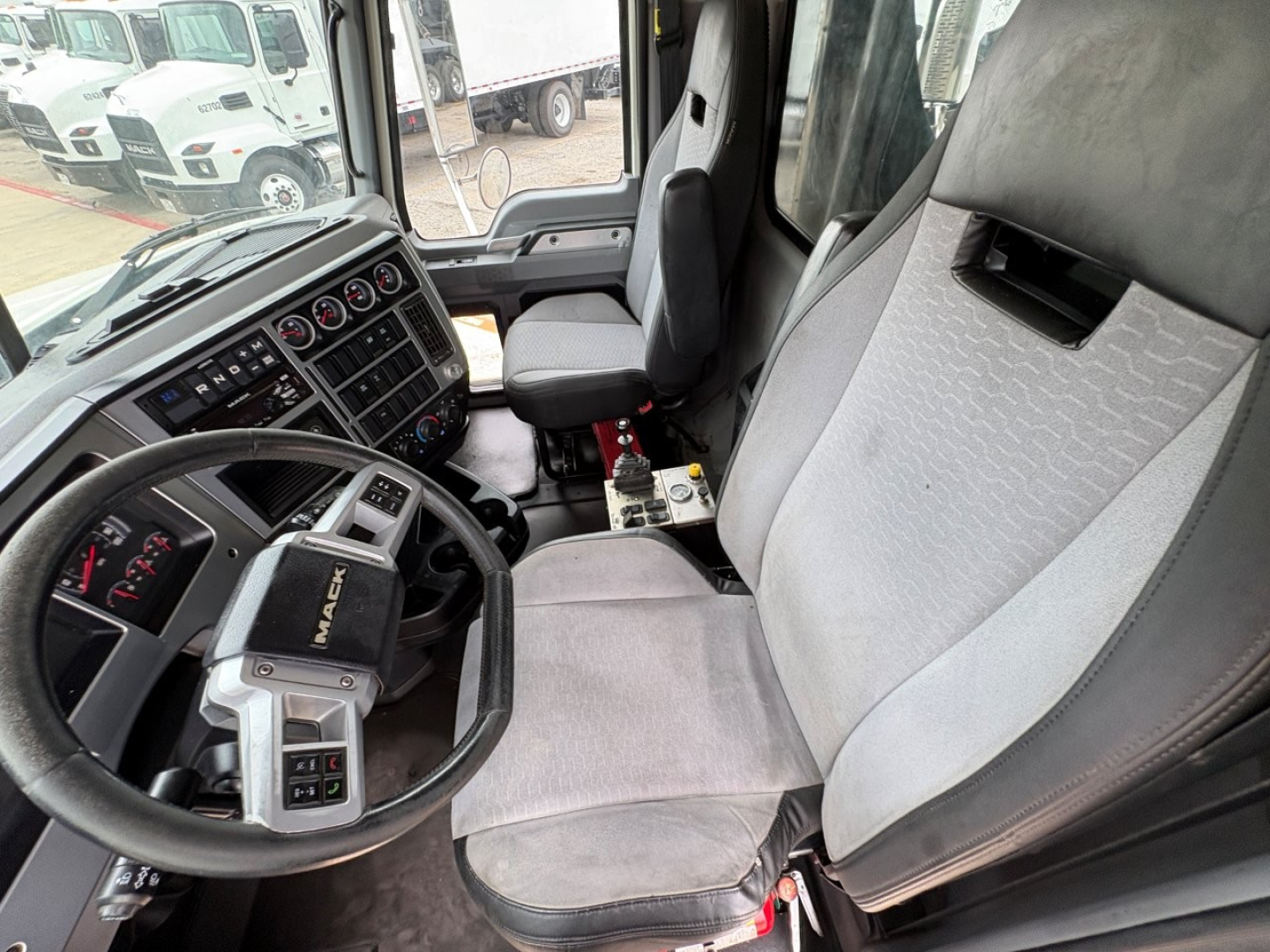 Truck cab interior of a 2021 Mack GR64F showing steering wheel, dashboard with gauges, and driver seat with light gray fabric. Visible control panel and center console.