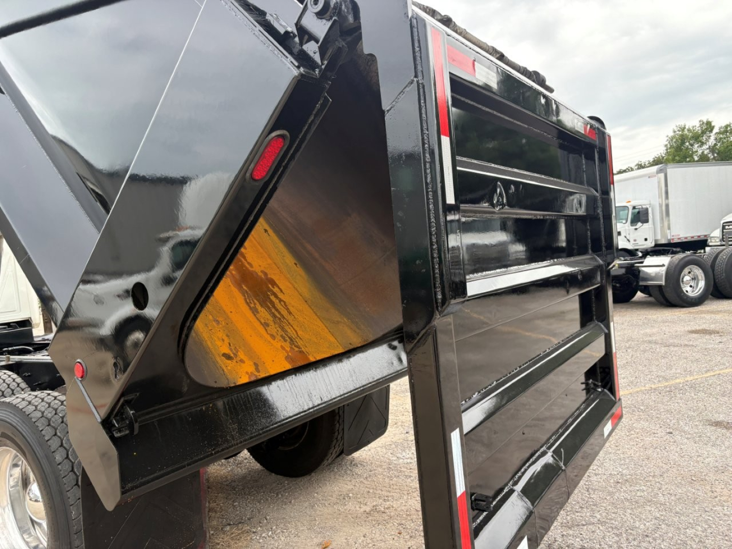 Dump truck body side with black steel panels and orange reflective strip, hydraulic dump bed raised, Mack GR64F truck in background.
