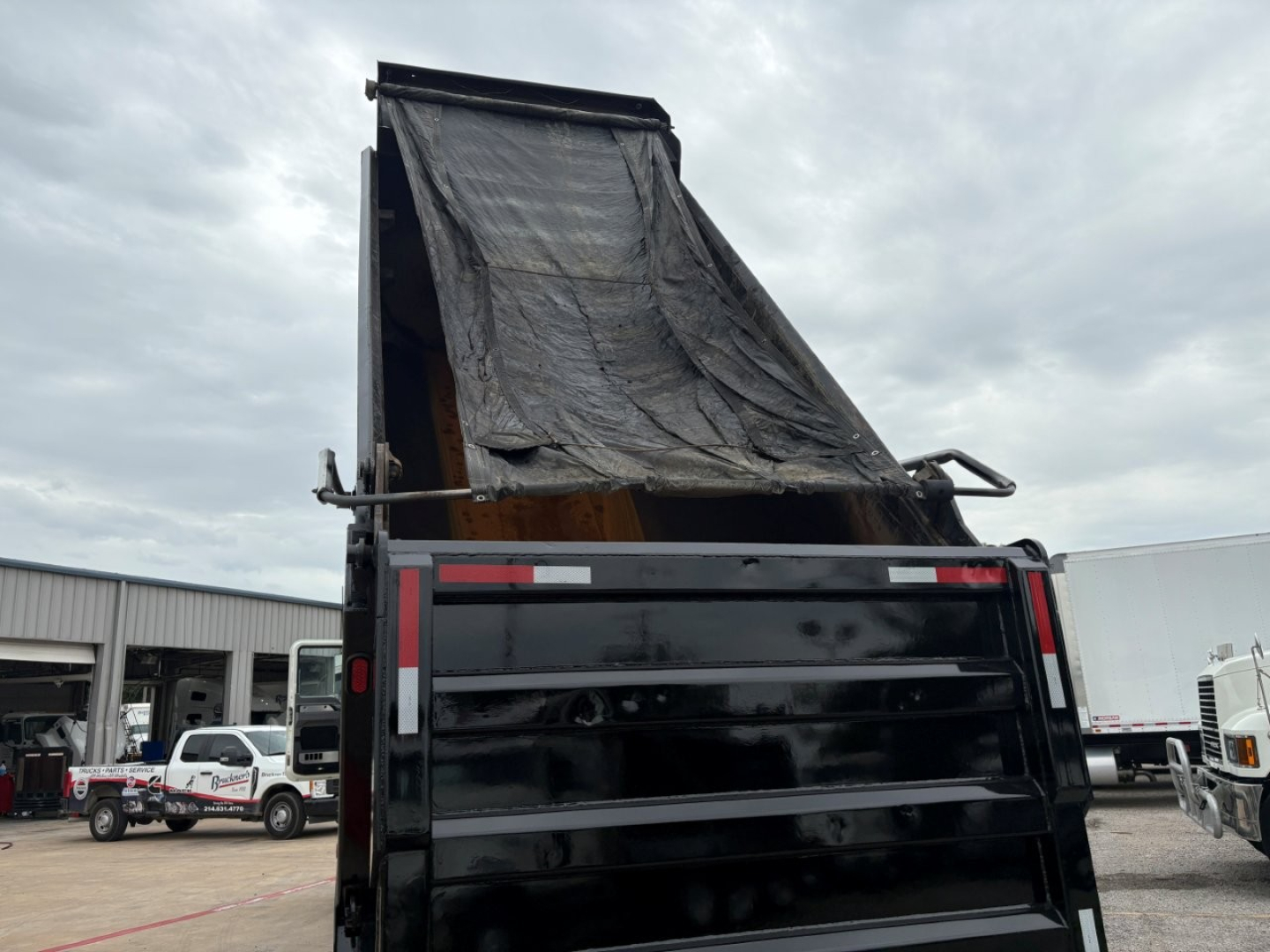 Dump truck bed raised with black dump body, tarp cover visible, side panels and rear tailgate, cloudy sky.