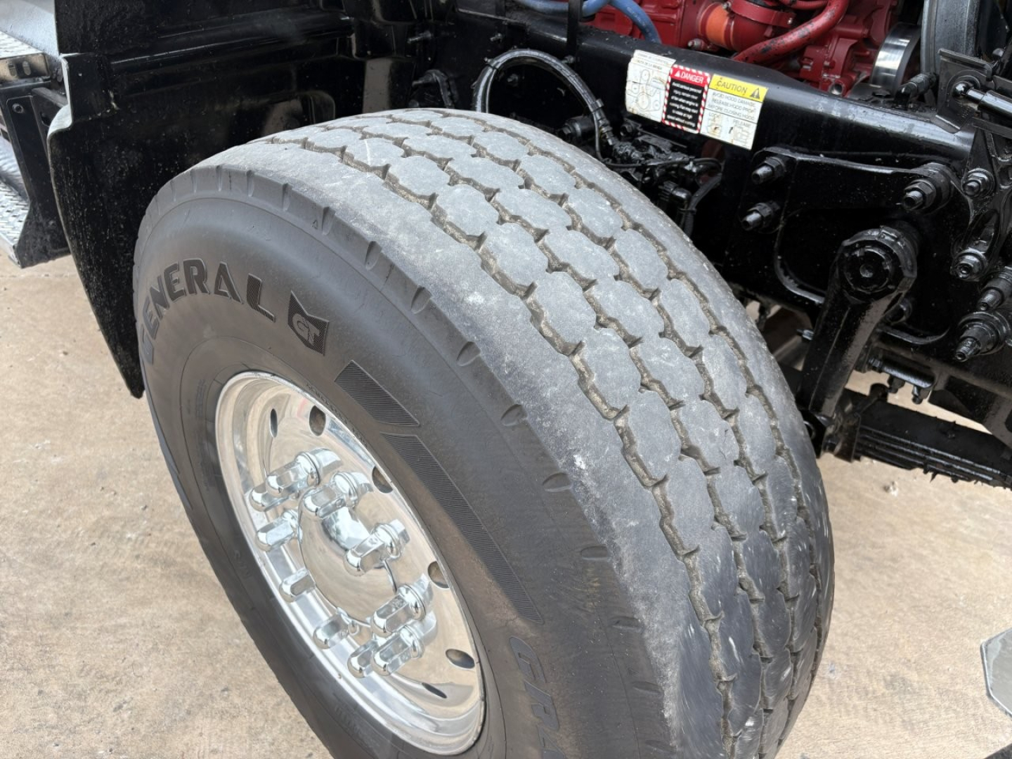 Front left wheel and tire of a 2021 Mack GR64F dump truck showing General tire and chrome wheel, part of the suspension and engine bay area visible.