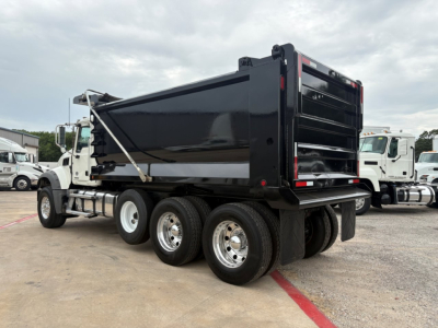 Dump truck chassis with black dump bed and triple rear axles, Mack GR64F, side view showing wheels and hydraulic bed hinges.