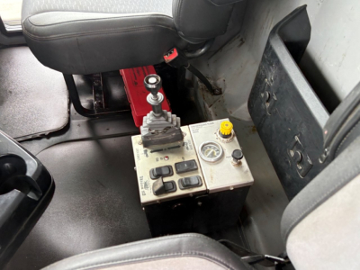 Cab interior of a Mack GR64F truck showing a joystick-style control lever and a metal control panel with gauges and buttons mounted near the driver seat.