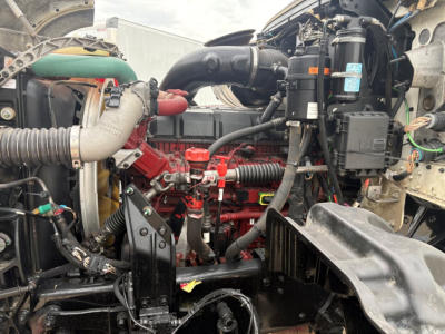 Truck engine bay showing Mack GR64F diesel engine with red engine block, hoses, and various components including coolant hoses and air intake.