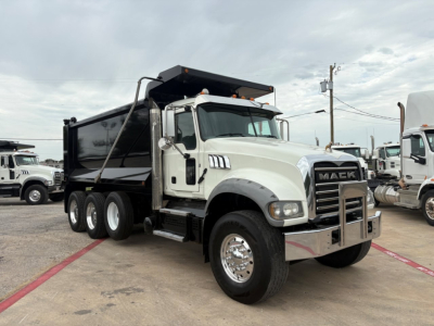 Dump truck showing a 2021 Mack GR64F with a white cab and black dump bed, visible front grille with MACK logo, large side mirrors, and chrome bumper.