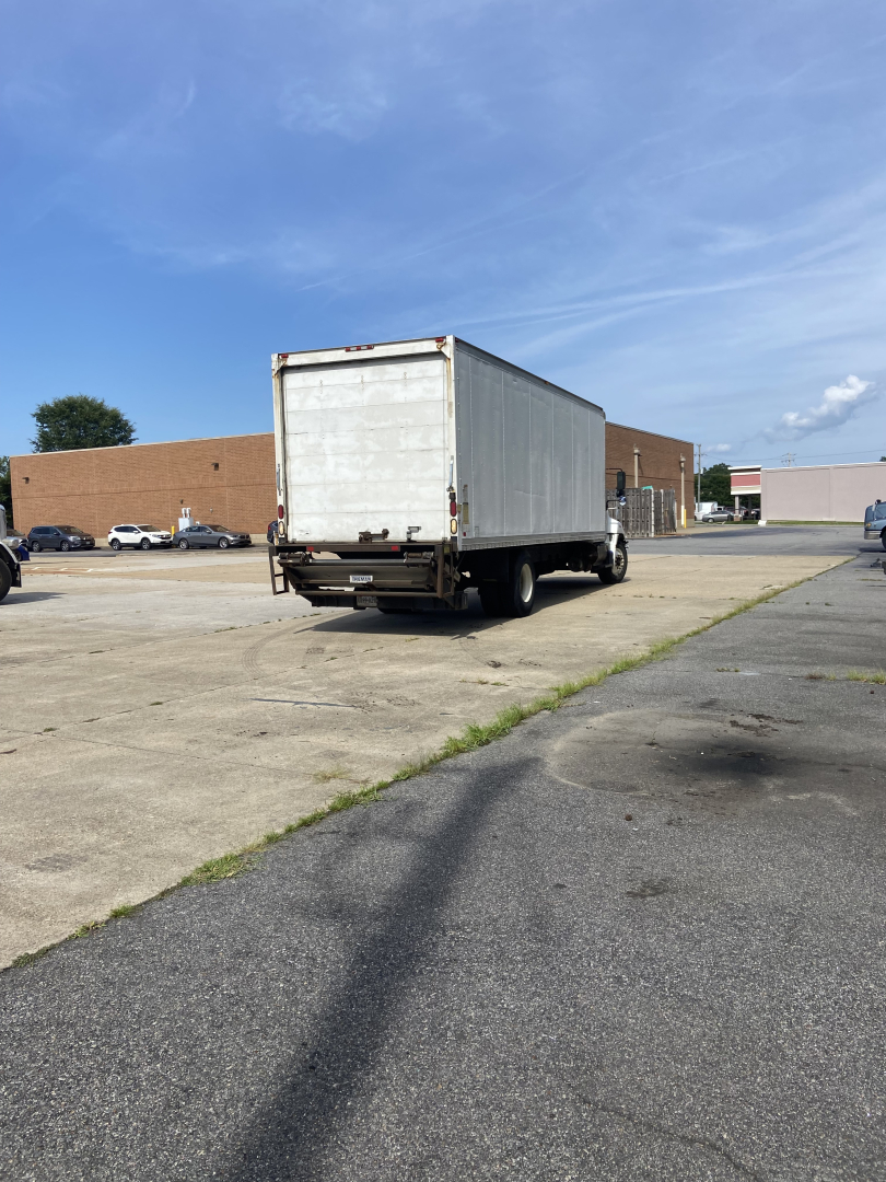 Rear view of a white box truck trailer attached to a cab, on a paved lot with a brick building in the background.