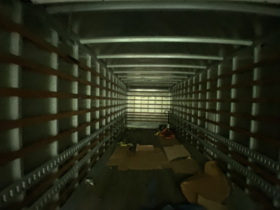 Interior of a truck cargo area with metal sidewalls and floor, dim lighting, scattered cardboard and debris along the floor.