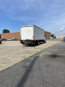 Rear view of a white box truck trailer attached to a cab, on a paved lot with a brick building in the background.