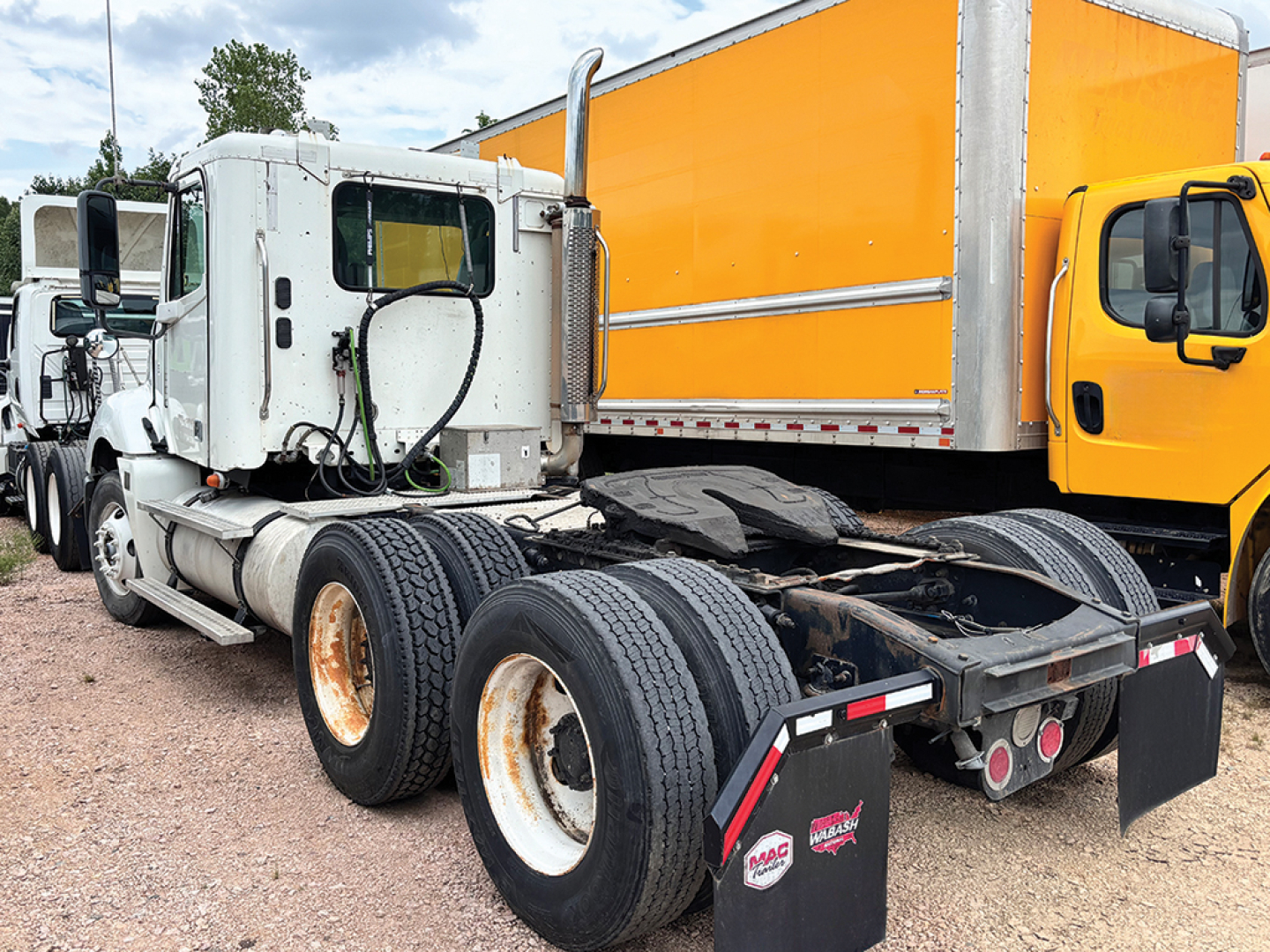 Truck chassis and cab of a white Freightliner CL120 Columbia tractor showing rear twin axles and fifth-wheel area with black tires and orange box truck in background