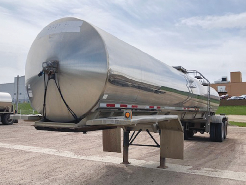 Stainless steel tanker trailer mounted on semi-truck chassis with cylindrical tank, support legs down, rear view showing piping and ladder, reflective finish.