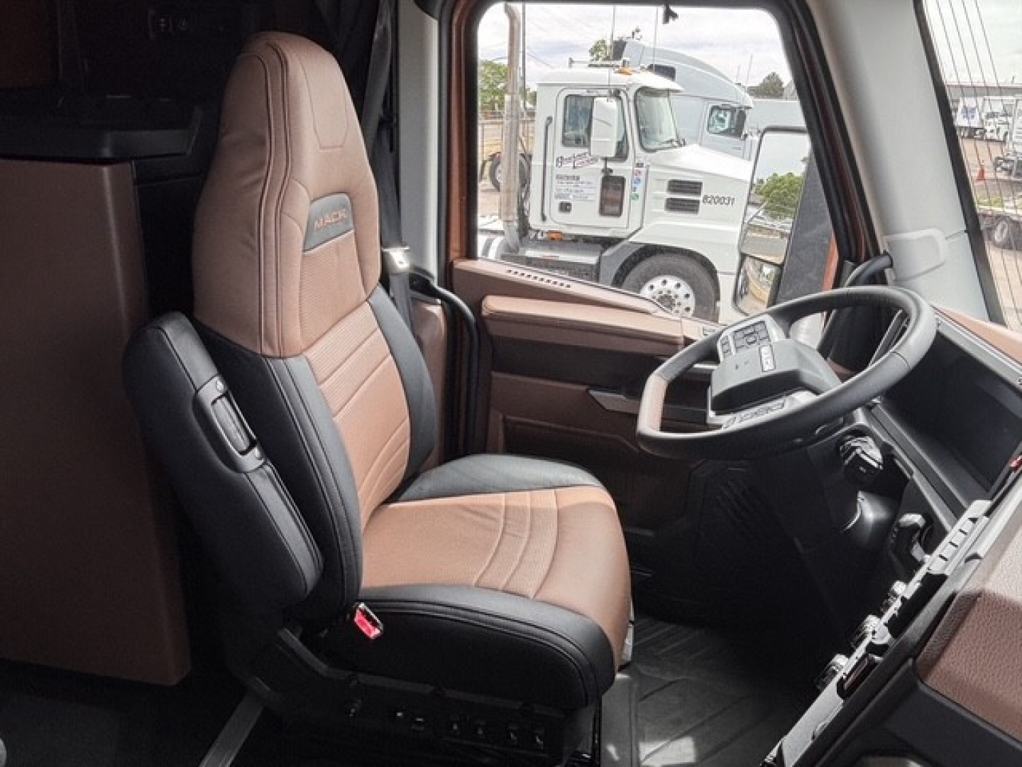 Driver seat and dashboard of a new 2027 Mack PR64T76H truck cabin with brown and black interior, steering wheel, and view through the windshield.