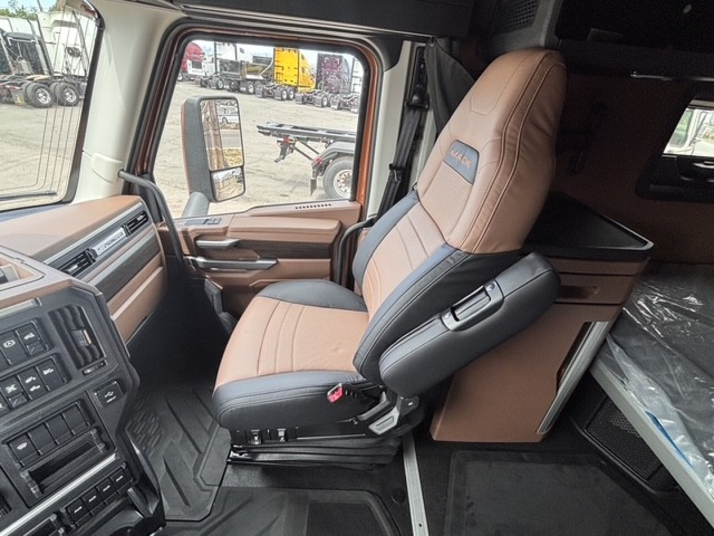 Truck cab interior showing tan and black driver seat with armrest, steering area, and sleeper compartment behind. CAB view from left side door.
