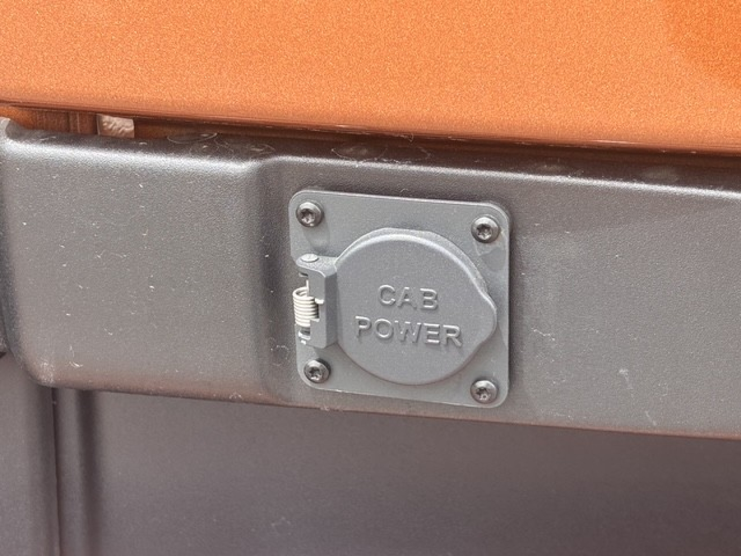 Tractor-trailer truck engine bay area showing a round power take-off (CAB POWER) cover on a gray metal panel; orange cab exterior above and black lower body.