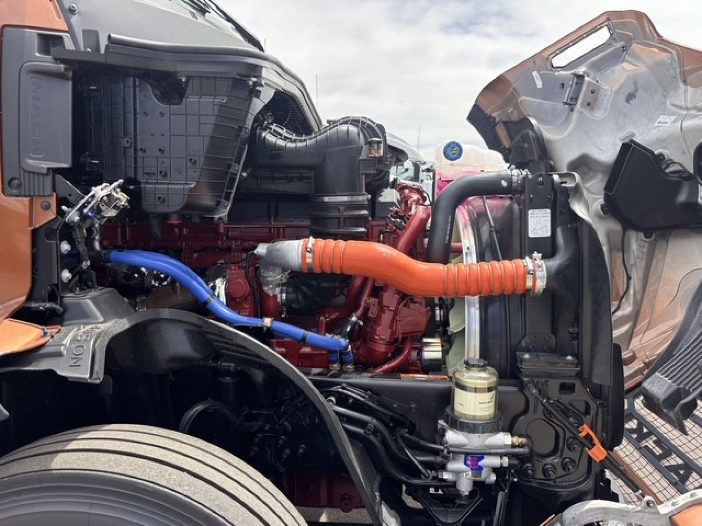 Truck engine bay of a 2027 Mack PR64T76H with orange and blue hoses, red engine block, exposed components, and open hood.