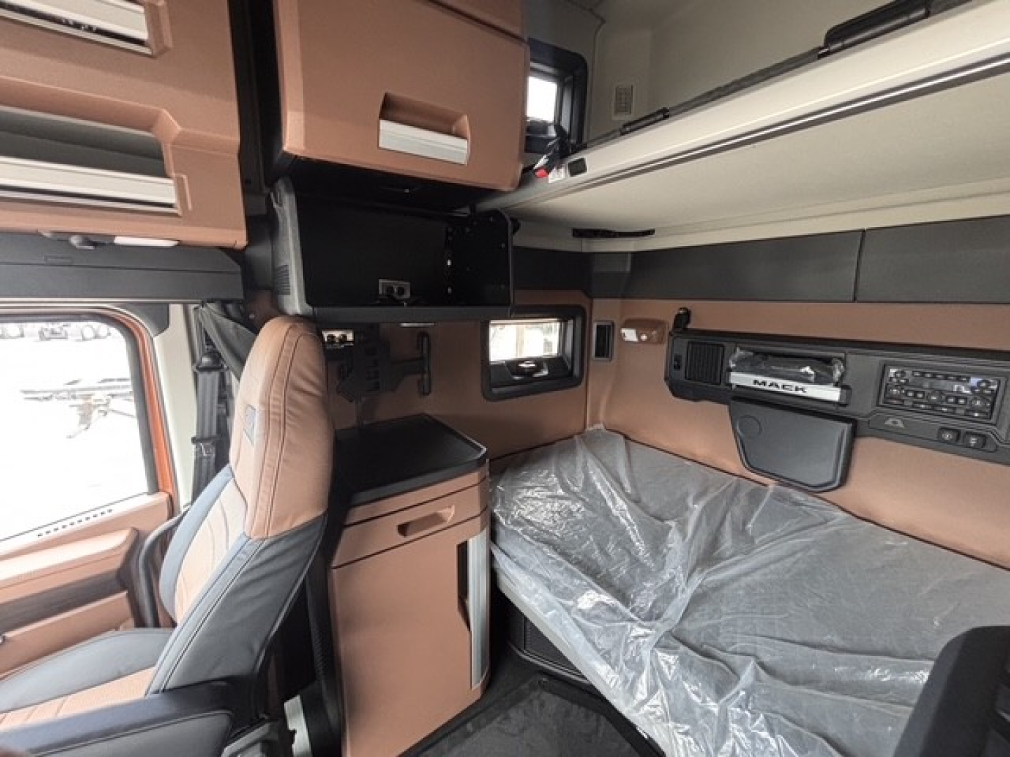 Interior view of a new 2027 Mack PR64T76H sleeper cab showing tan and gray dashboard, seating, and storage compartments with bed area covered by a protective sheet.
