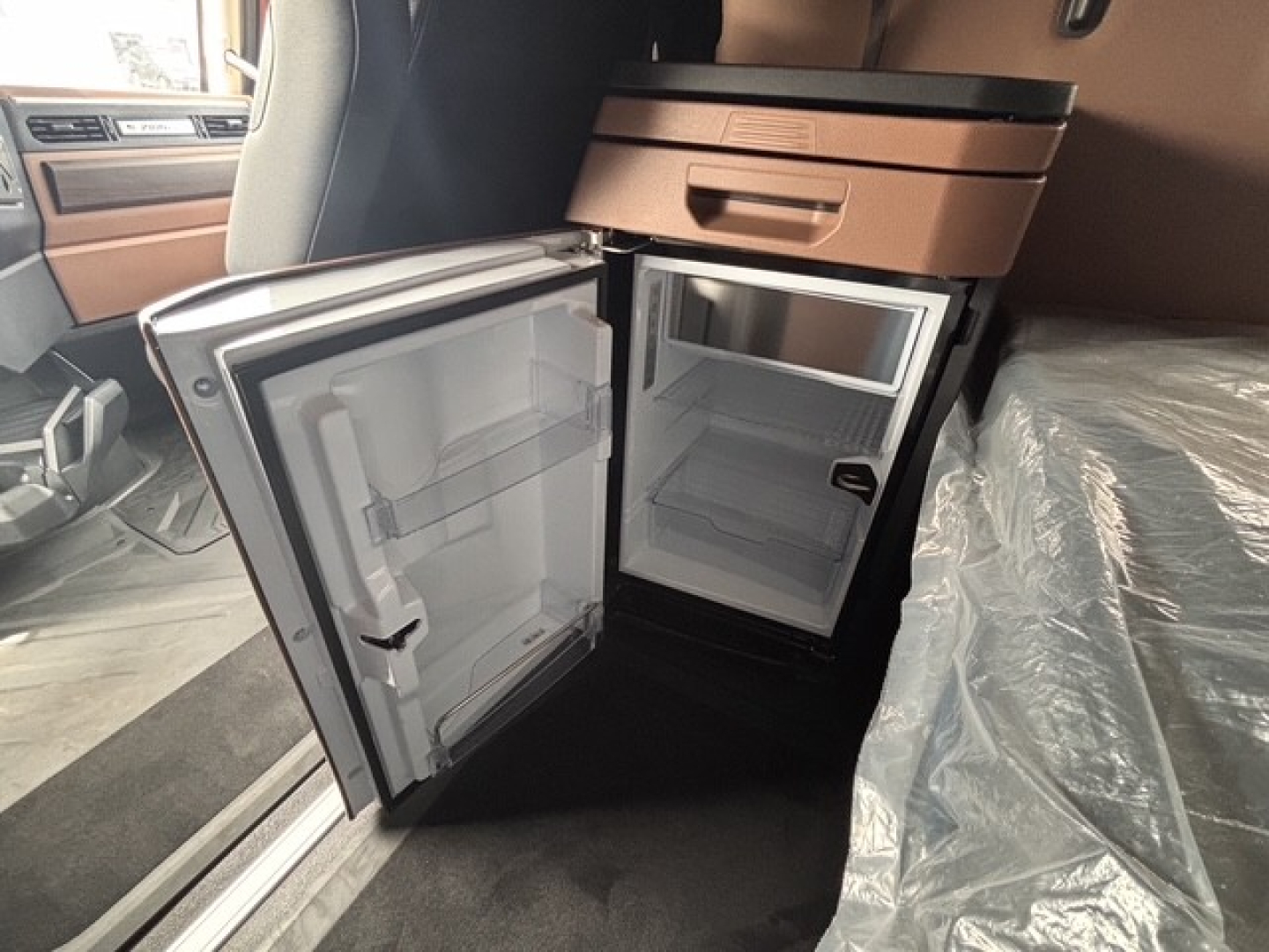 Truck cab interior showing open refrigerator/freezer in a Mack PR64T76H sleeper. Includes compact fridge with empty shelves and freezer compartment.