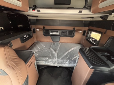Truck cab interior showing dashboard with central control panel and brown/tan upholstery; plastic cover on mattress area and overhead bunk storage visible.