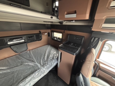 Interior of Mack PR64T76H sleeper cab showing bunk and control panel, kitchenette area with small fridge and storage compartments.