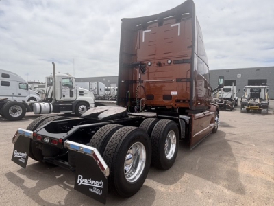 Truck rear/side view showing fifth wheel, dual rear axles, and mud flaps in a lot of new Mack PR64T76H tractor.