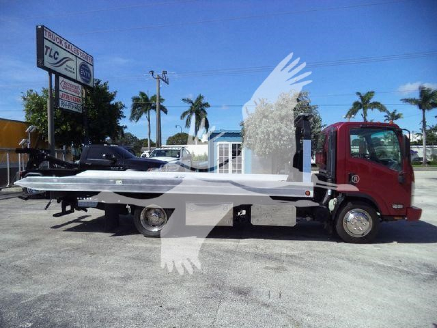 Isuzu NPR XD rollback truck with a flatbed, side view showing truck cab and bed. Signage in background reads TLC and truck sales. No clear model-specific text on the vehicle.