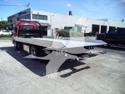 Isuzu NPR XD rollback truck bed with flatbed extending rear, viewed from left-rear; light gray concrete lot and building in background.