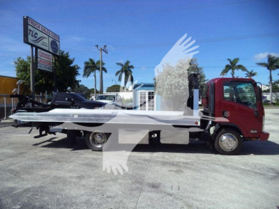 Isuzu NPR XD rollback truck with a flatbed, side view showing truck cab and bed. Signage in background reads TLC and truck sales. No clear model-specific text on the vehicle.