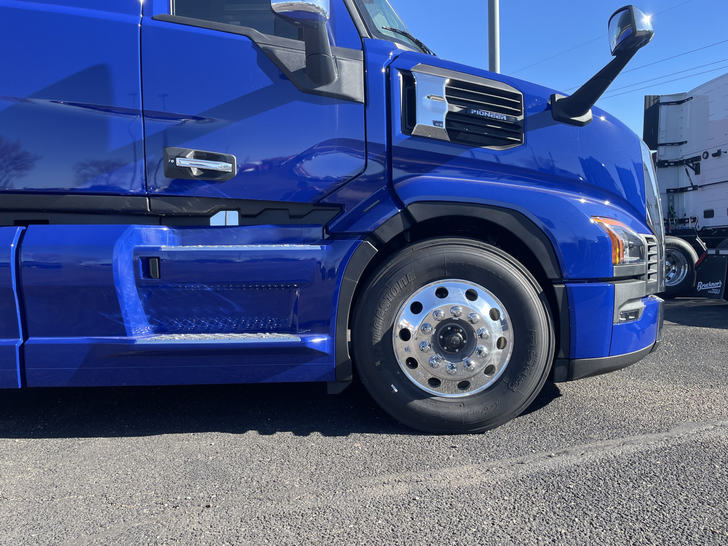 Blue Mack PR64T76H truck side front wheel well and cab details with chrome mirrors and badge, showing front wheel and lower fender.