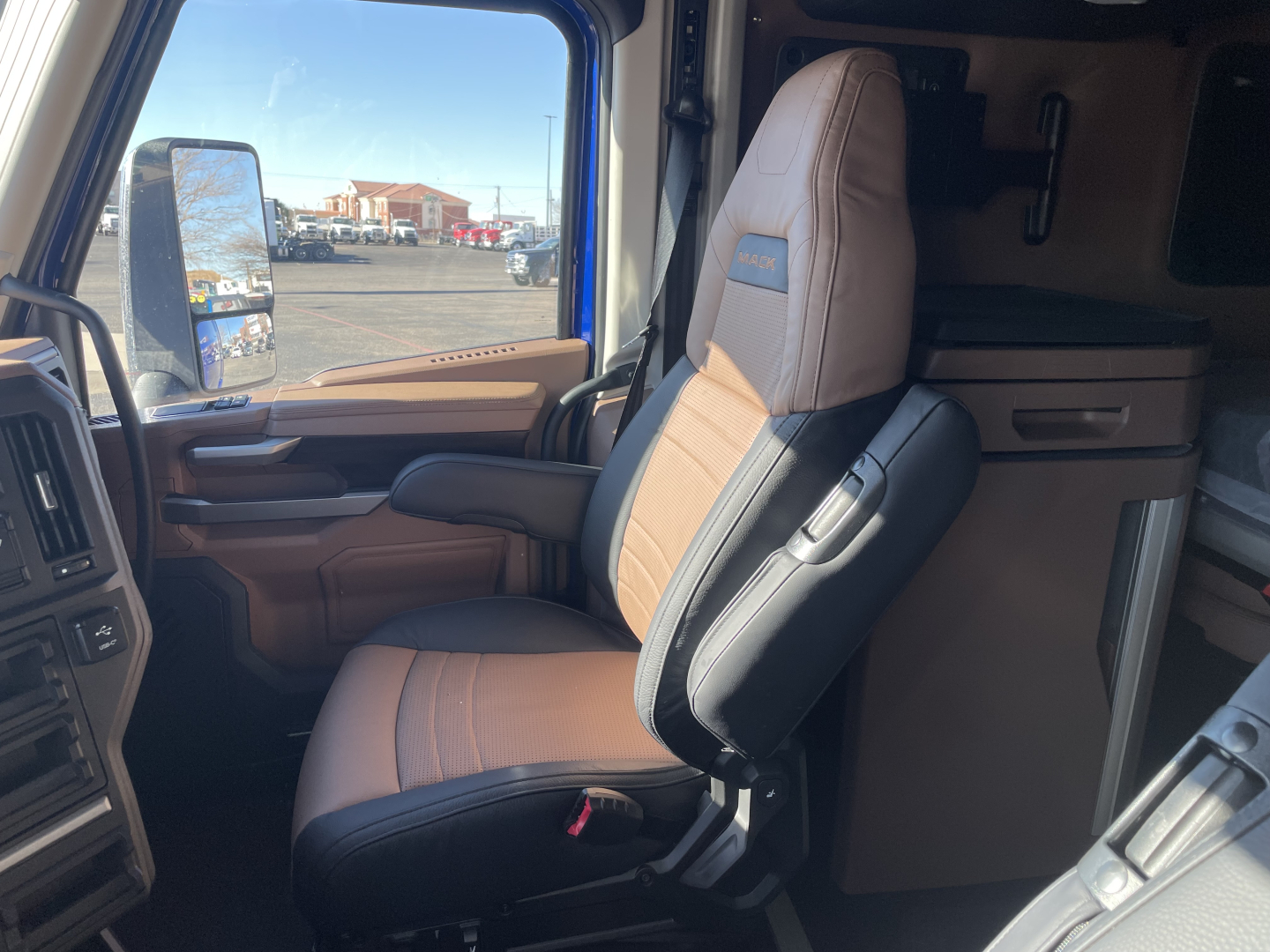 Driver seat area of a 2026 Mack PR64T76H truck interior showing tan and black seats, door panel, and part of the dashboard.